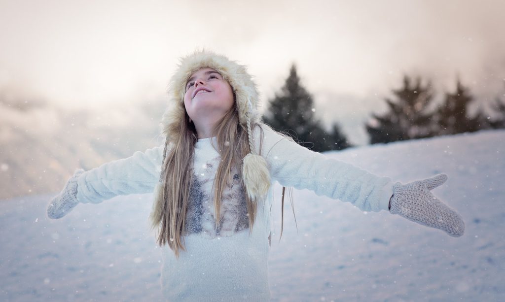 Menina olhando para o céu de braços abertos a espera dos flocos de neve que caem sobre ela. Ela está de luvas, gorro e casaco apropriados para a neve.